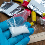 Blue-gloved hand holding bag of white powder, other substances nearby.
