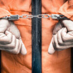 Handcuffed person in orange jumpsuit behind prison bars.